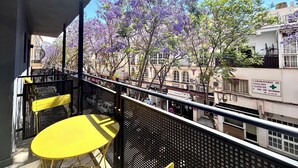 A balcony with yellow chairs and a table, overlooking a street with buildings and purple flowering trees. - ZUR Boutique Hostel (Fuengirola)