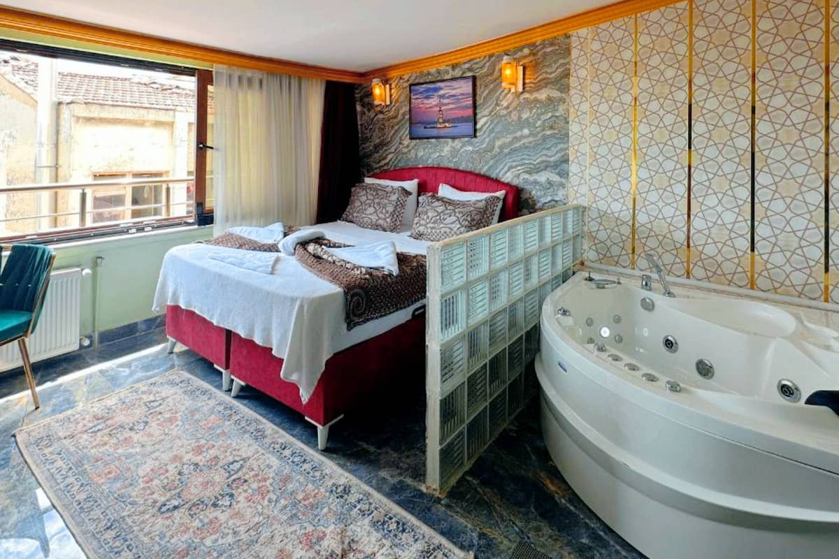 A bedroom with a bed, a large window, a view of the outside, a wall-mounted TV, and a bathtub.