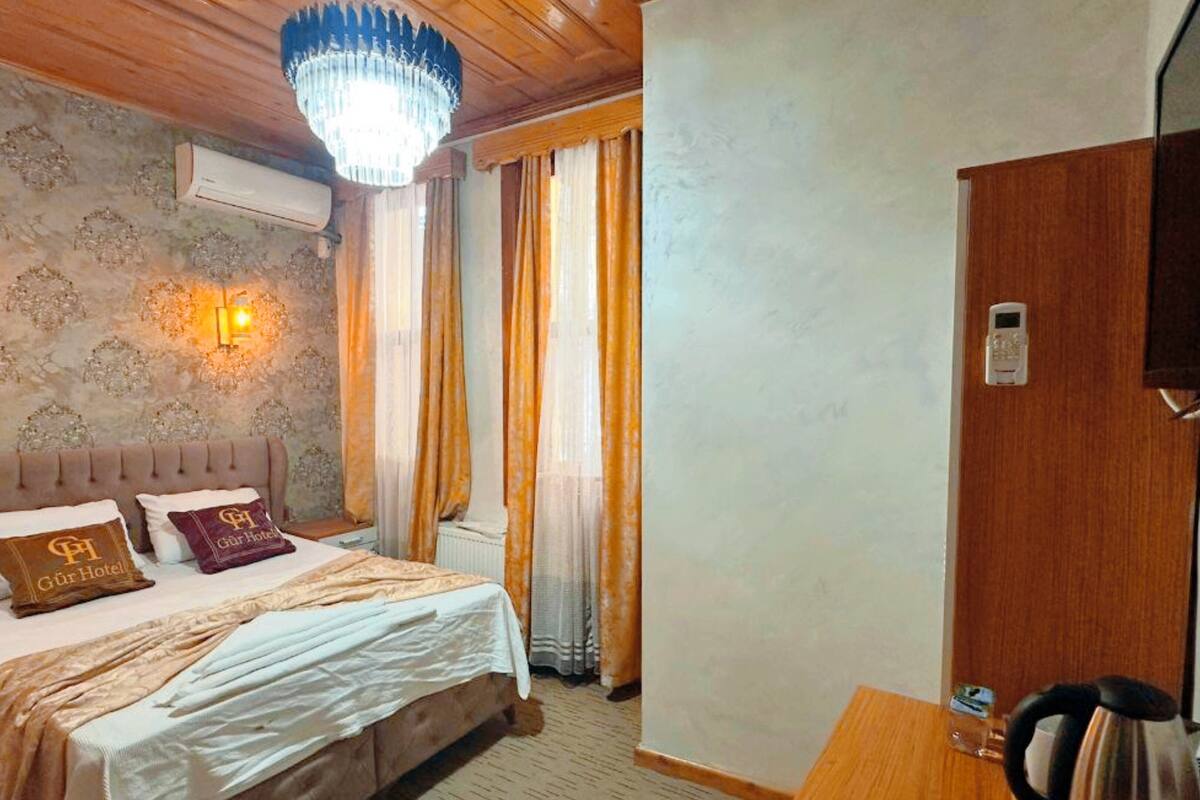 A hotel room with a bed, a chandelier, a wall-mounted air conditioner, a wooden desk with a kettle, and a door with a lock.