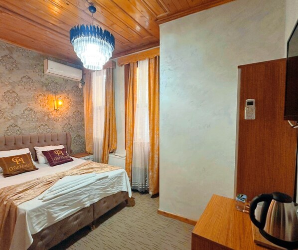 Una habitación de hotel con cama, candelabro, aire acondicionado instalado en la pared, escritorio de madera con tetera y puerta con cerradura.