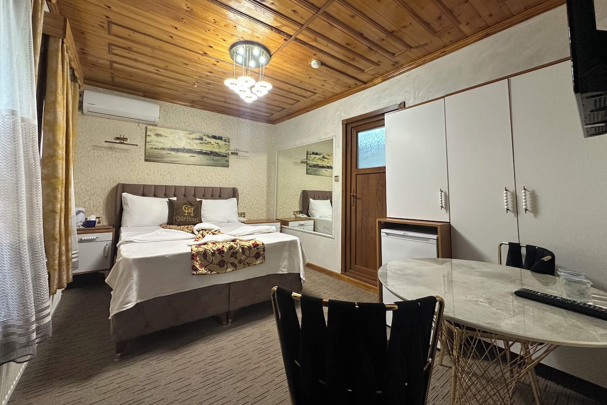 A hotel room with a wooden ceiling, a bed with a headboard, a bedside table, a mirror, a wardrobe, a dining table with chairs, and a small refrigerator.