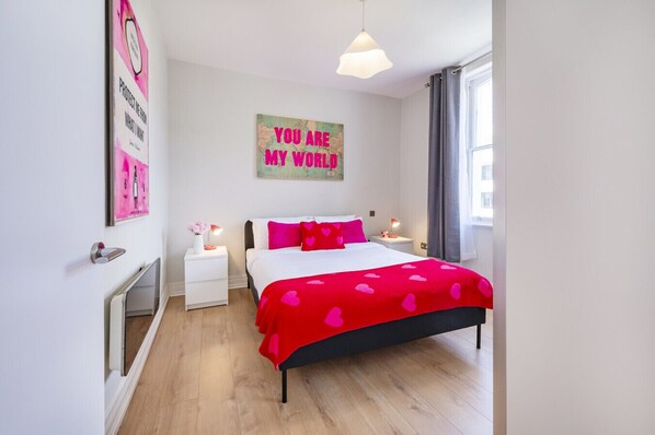 A bedroom with a bed, two bedside tables, a radiator, a window with curtains, and a wall with a motivational poster.
