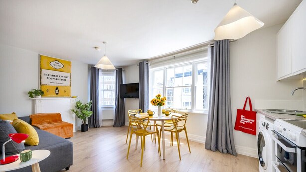 A modern living room with a sofa, a dining table with chairs, a TV, and a kitchen area with a washing machine.