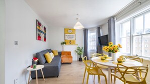 A modern living room with a grey sofa, yellow chairs, a small table, and a dining table with yellow chairs. There is a TV on the wall and a window with curtains.