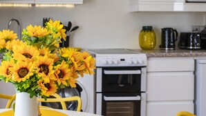 A modern kitchen with white cabinets, a white stove, and a yellow dining table with a vase of sunflowers and pineapple-shaped mugs.