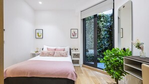 A modern bedroom with a large bed, a bedside table, a nightstand, a mirror, and a view of greenery outside.