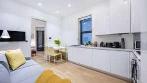 A modern kitchen with white cabinets, a dining area with a table and chairs, and a living room with a sofa and a yellow throw blanket.