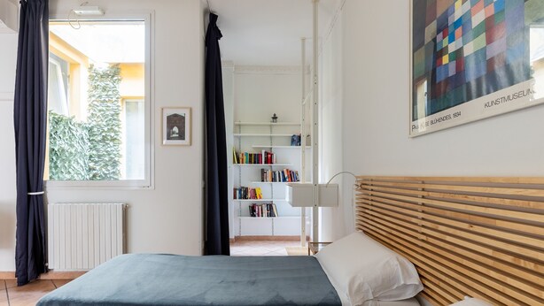 Un dormitorio con cama, mesita de noche, lámpara, estantería y ventana con cortinas.