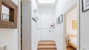 Un pasillo estrecho con suelo de baldosas, una puerta blanca al final y una cama con cabecero de madera en la habitación contigua.