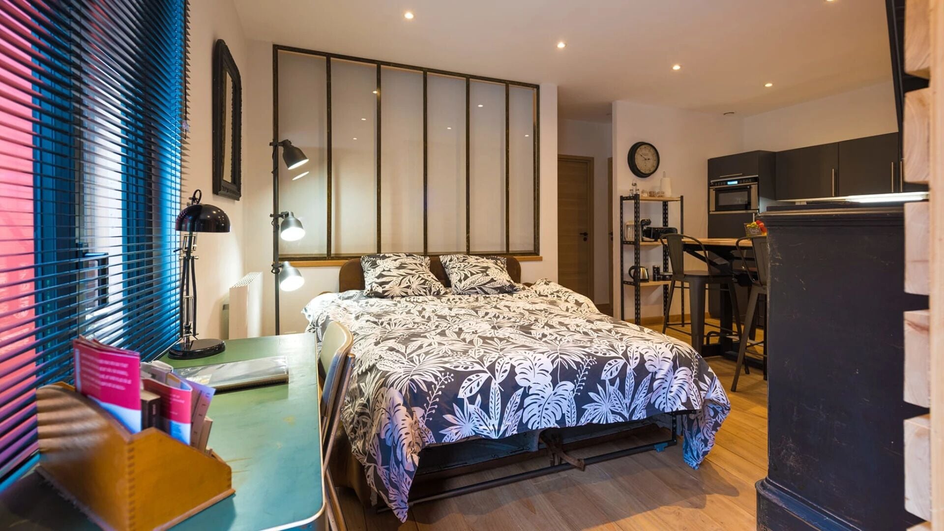 A bedroom with a bed, bedside lamps, a desk, a chair, a wardrobe, and a kitchen area with a refrigerator and microwave.