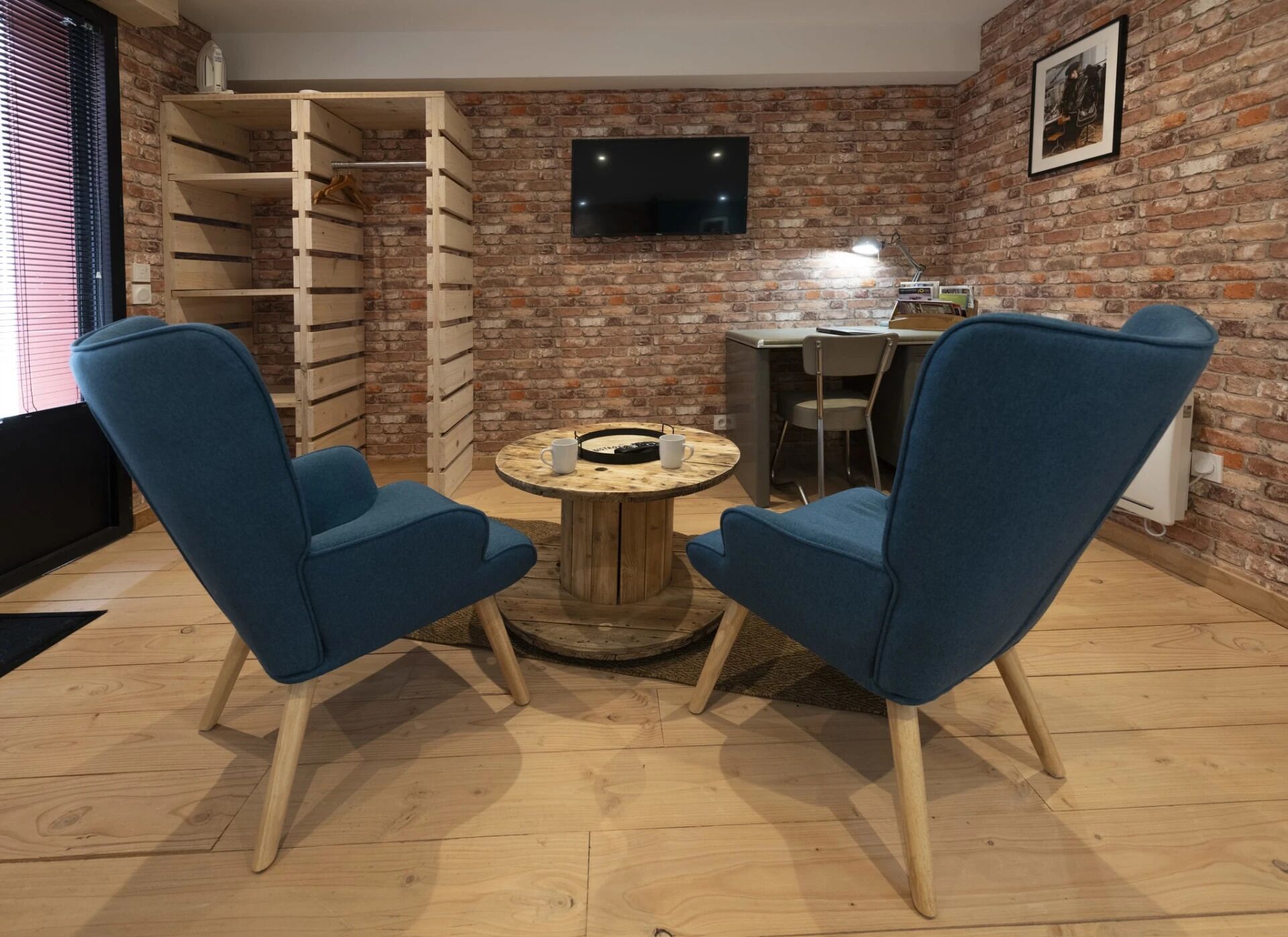 Una habitación con pared de ladrillo, un televisor montado sobre un escritorio, dos sillas azules y una mesa redonda de madera con una bandeja.