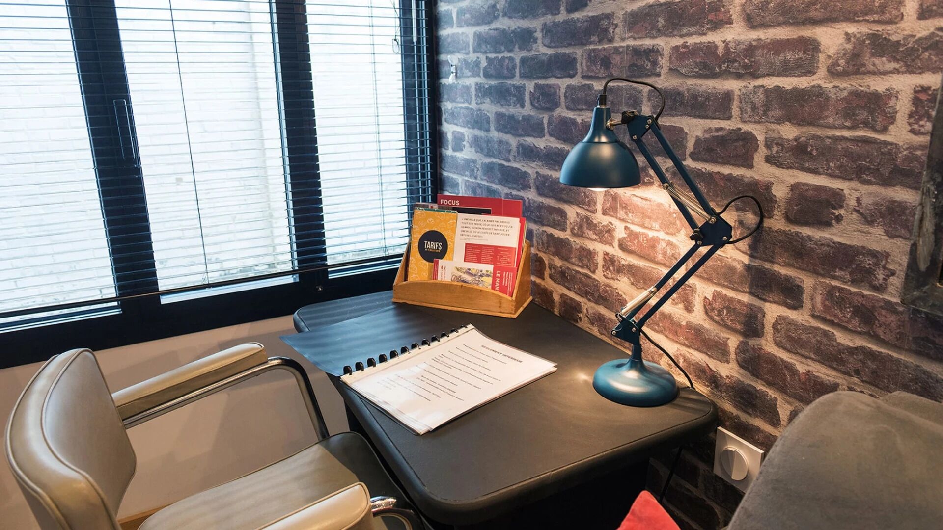 Ein Arbeitsplatz mit Schreibtisch, Stuhl und Lampe. Ein Fenster mit Jalousien, eine Ziegelwand und ein kleines Regal mit Gegenständen.