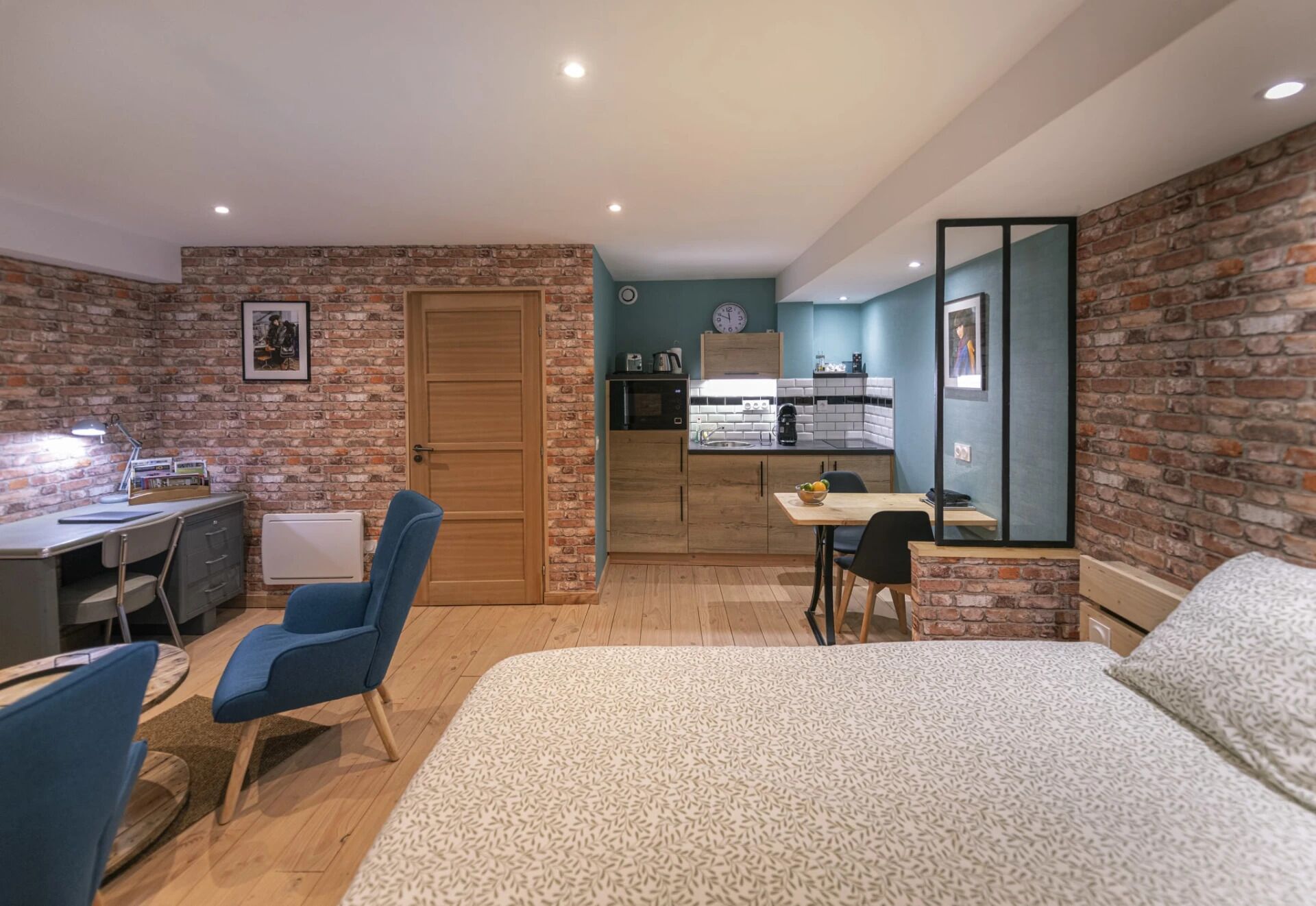 A compact room with a bed, a desk, a chair, a small kitchenette, and a door.