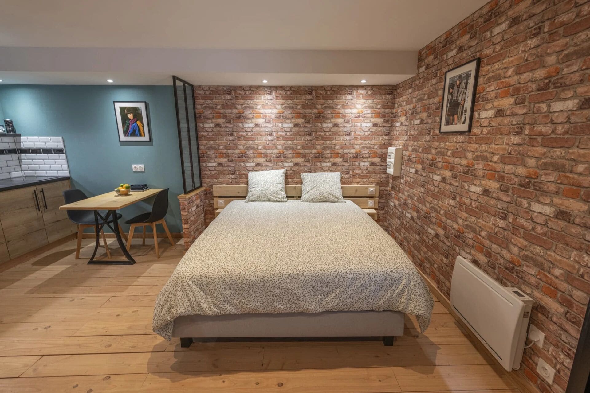 Un dormitorio con pared de ladrillo, una cama con colcha estampada, un escritorio con silla y una pequeña área de kitchenette.