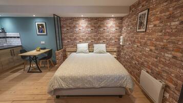 Un dormitorio con pared de ladrillo, una cama con colcha estampada, un escritorio con silla y una pequeña área de kitchenette.