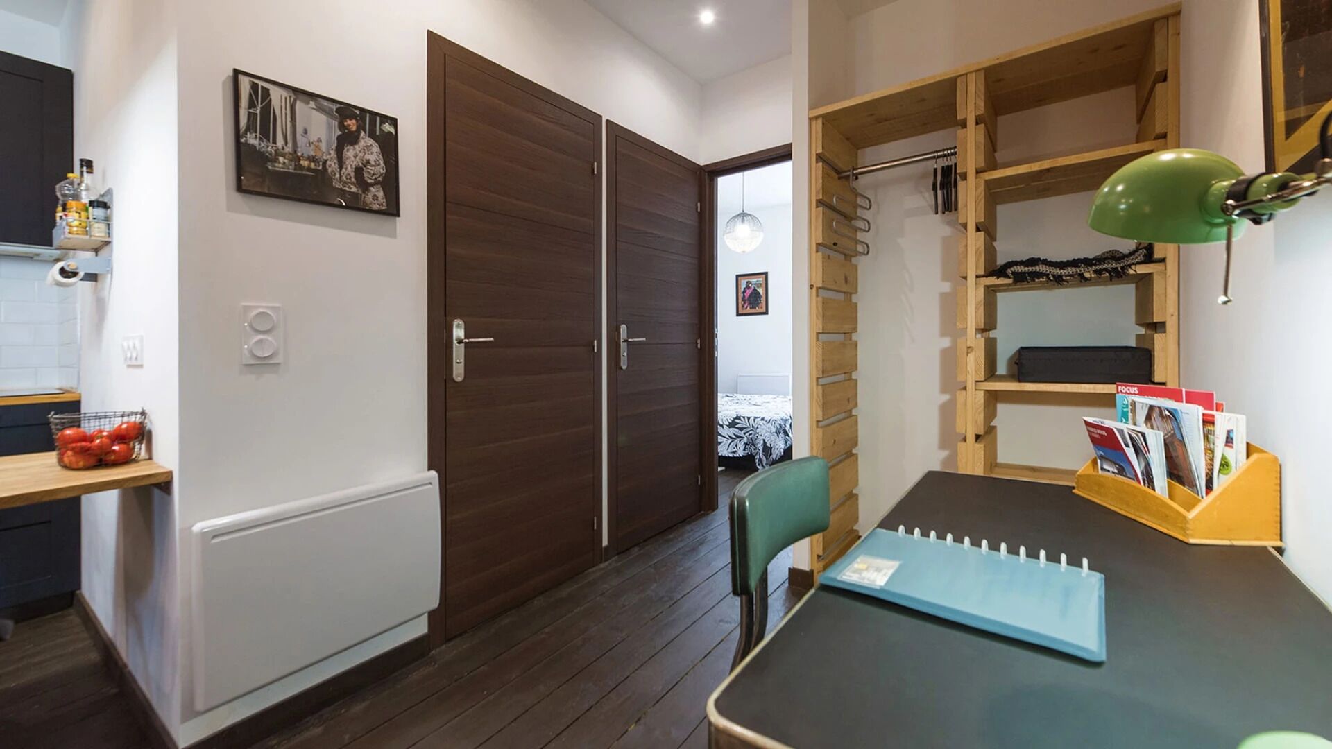 A compact room with a wooden wardrobe, a small table with a notebook, a green chair, and a wooden shelf with magazines.