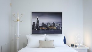 A neatly arranged bedroom with a cityscape view, a white bed, a bedside table with a lamp, and a wall-mounted light fixture. - Escape Villa in Melissochori (Oraiokastro)
