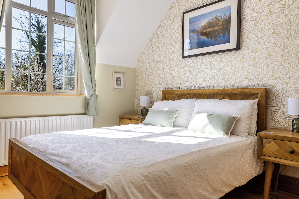 Een net opgemaakt bed met een bloemenbedekker, een nachtkastje met een lamp en een raam met gordijnen.