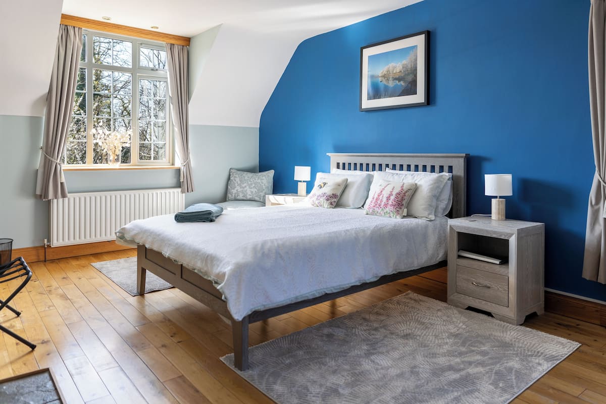 Een slaapkamer met een bed, nachtkastjes, een raam met gordijnen, een radiator en een schilderij aan de muur.