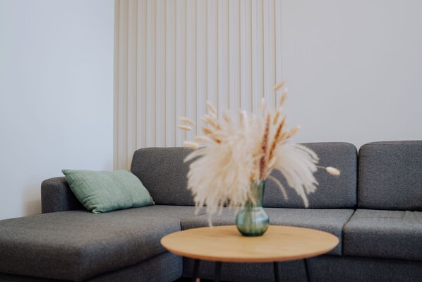 A grey sofa with a green pillow, a round wooden table, and a vase with dried pampas grass.