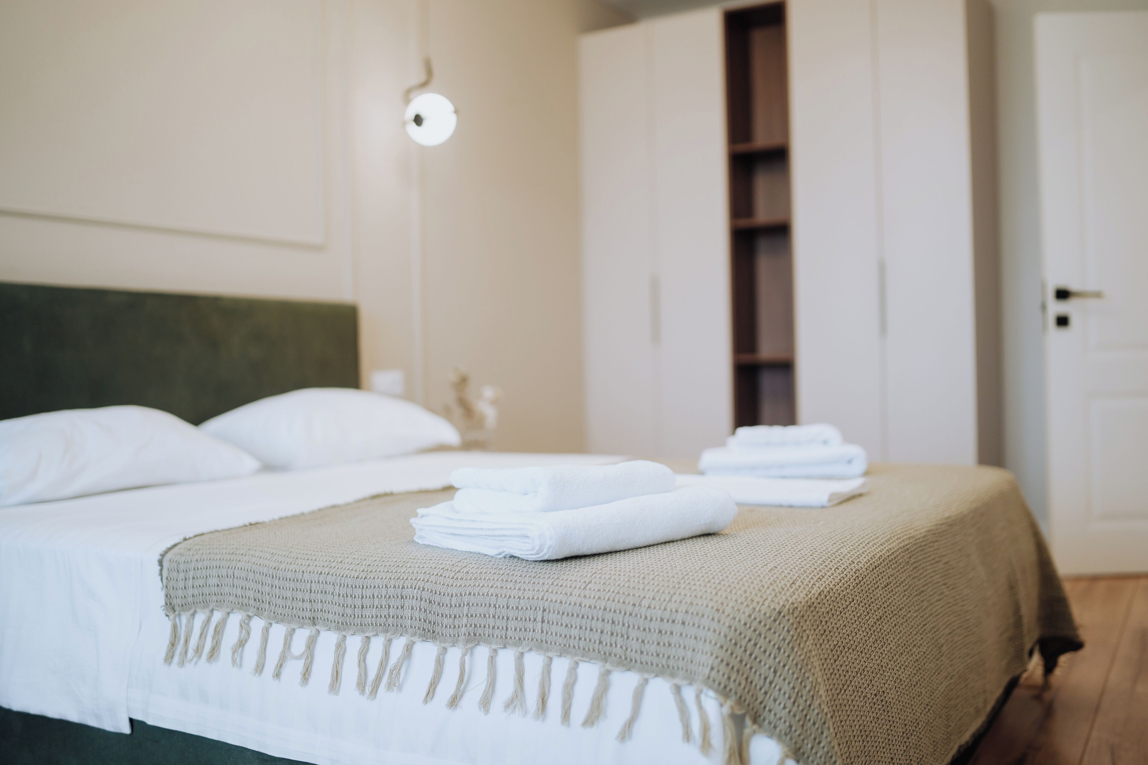 A neatly made bed with white linens and a beige throw blanket, a wall-mounted light fixture, and a door to a closet.