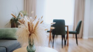 Une table ronde en bois, avec un vase rempli de plumes d’herbe des pampas au premier plan. À l’arrière-plan, on aperçoit un canapé vert et un coin repas avec une chaise.