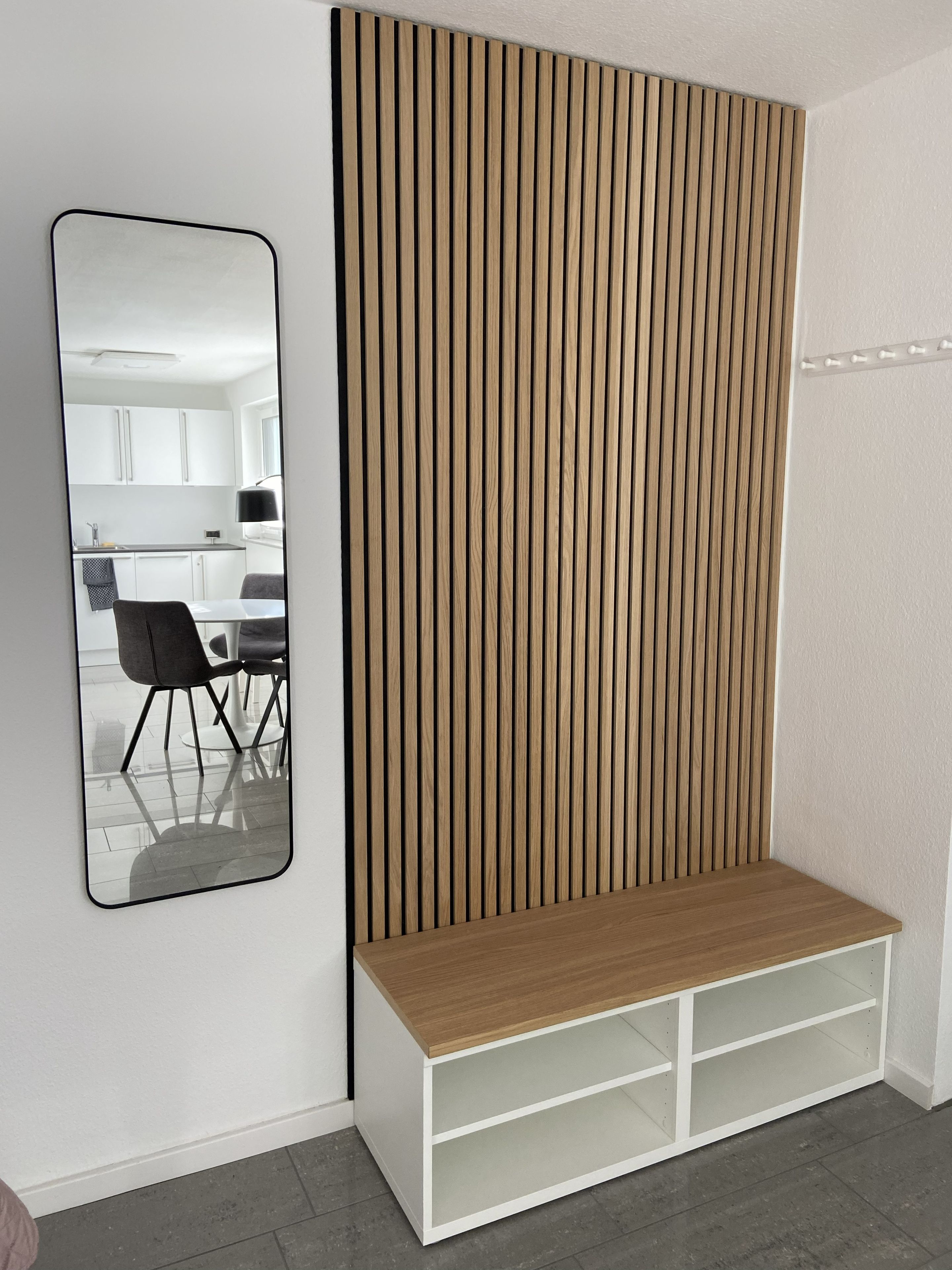 A room with a white wall, a wooden slatted partition, a white bench with shelves, and a mirror reflecting a kitchen area.
