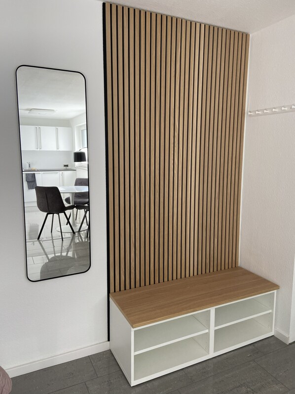 A room with a white wall, a wooden slatted partition, a white bench with shelves, and a mirror reflecting a kitchen area. - New vacation apartment in a prime location—close to the lake and in a quiet area (Überlingen)