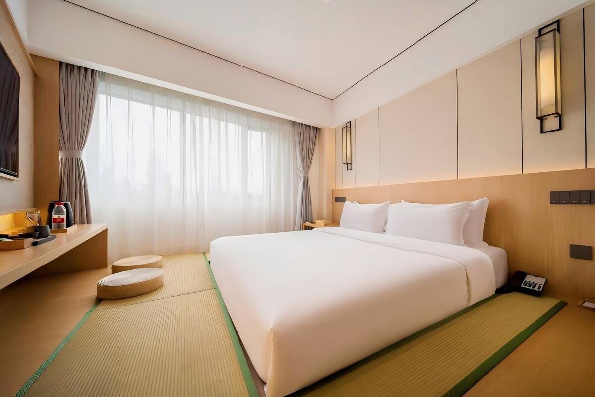 Ein Hotelzimmer mit einem großen Bett, einem Nachttisch mit Lampe, einem Fernseher und einem Fenster mit durchsichtigen Vorhängen.