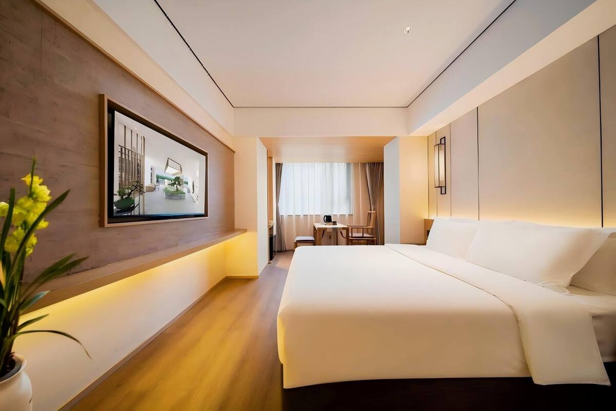 Ein modernes Hotelzimmer mit einem großen Bett, einem Schreibtisch mit Stuhl, einer Zimmerpflanze und einem gerahmten Bild an der Wand.