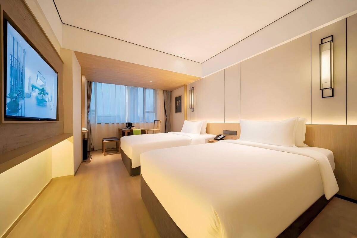 Ein Hotelzimmer mit zwei Betten, einem Fernseher, einem Schreibtisch mit Stuhl und einem Fenster mit Vorhängen.