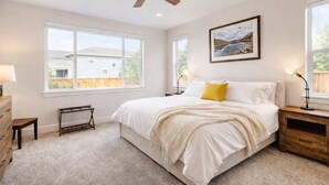 Quarto com cama grande, duas mesas de cabeceira, ventoinha no teto e janela com vista para uma casa.