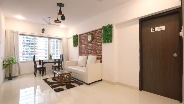 A modern living room with a sofa, coffee table, and dining area. A brick accent wall with a clock and greenery is visible. - 1 BHK Private Suite ( Gagal Home) (Mumbai)