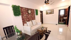 A modern living room with a brick accent wall, a sofa, a glass coffee table, and a dining area with a glass table and chairs. - 1 BHK Private Suite ( Gagal Home) (Mumbai)