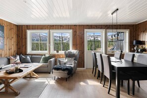 Una habitación con paneles de madera, un sofá, una silla, una mesa de comedor con sillas y ventanas amplias con vistas a montañas.