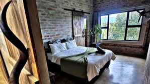 Ein rustikales Schlafzimmer mit Backsteinwand, Holzbalkendecke, einem Bett mit weißen Leinentüchern, einer Pflanze und einem Fenster mit Blick auf Bäume.