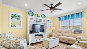A living room with a beige sofa, a floral-patterned couch, a television on a white cabinet, and a ceiling fan.