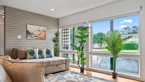 A modern living room with a brick wall, a large window, and a view of a building and greenery outside. - Relax in Style – 5BR Home w/ Pool, Hot Tub & Stunning Harbor Views (Galveston)