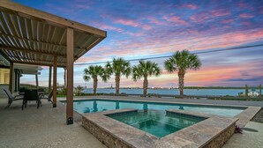 A swimming pool with a stone deck, a covered seating area with chairs, and palm trees. - Relax in Style – 5BR Home w/ Pool, Hot Tub & Stunning Harbor Views (Galveston)