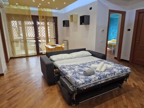 Ein Schlafzimmer mit einem Bett, einer Couch, einem kleinen Tisch und einem Stuhl. Eine Schiebetür führt zu einem Außenbereich.