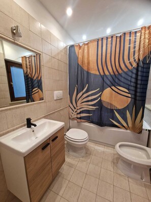 Ein Badezimmer mit Waschbecken, Toilette und Bidet, einer Duschvorhang mit Blättermotiv und einem Spiegel über dem Waschbecken.