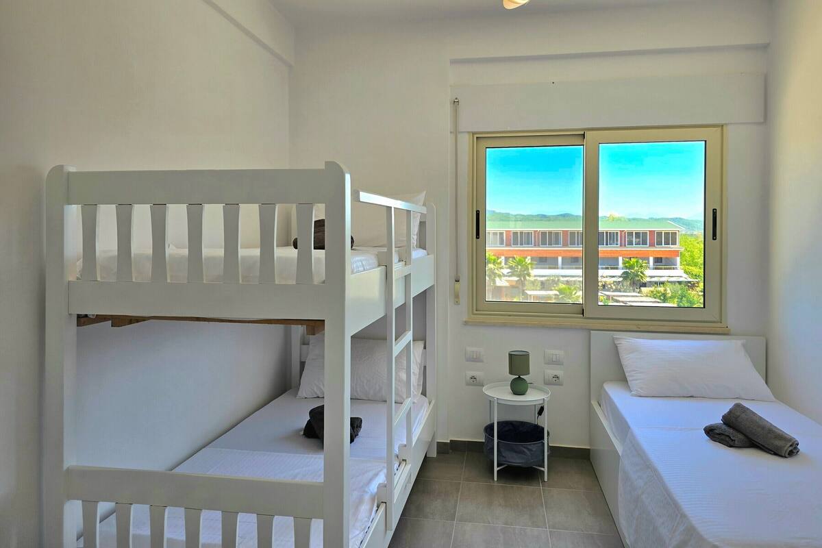 A room with bunk beds, a small table with a lamp, and a window with a view of buildings.