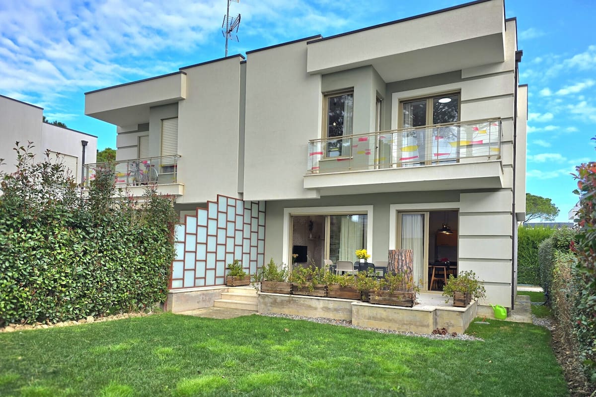 A modern two-story building with a balcony, a small garden area with plants, and a clear sky.