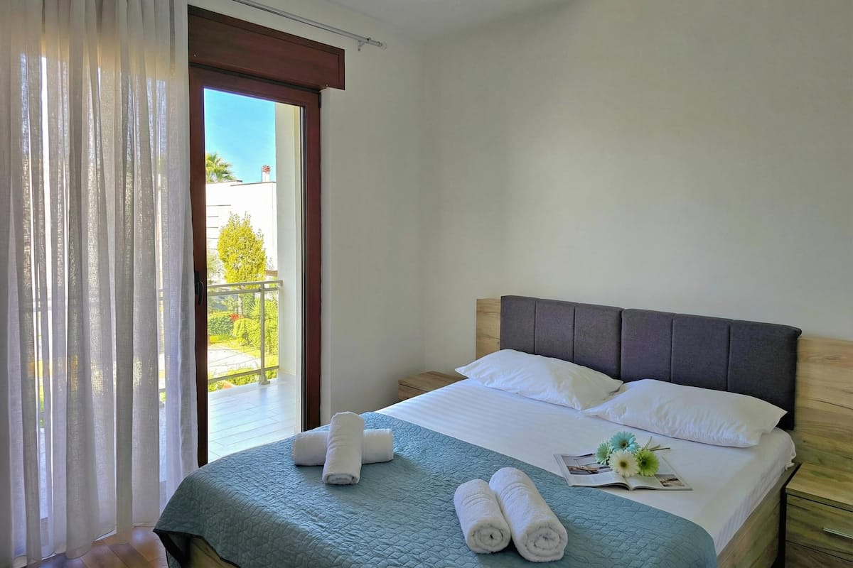 A hotel room with a bed, a nightstand, a window with curtains, and a view of greenery outside.