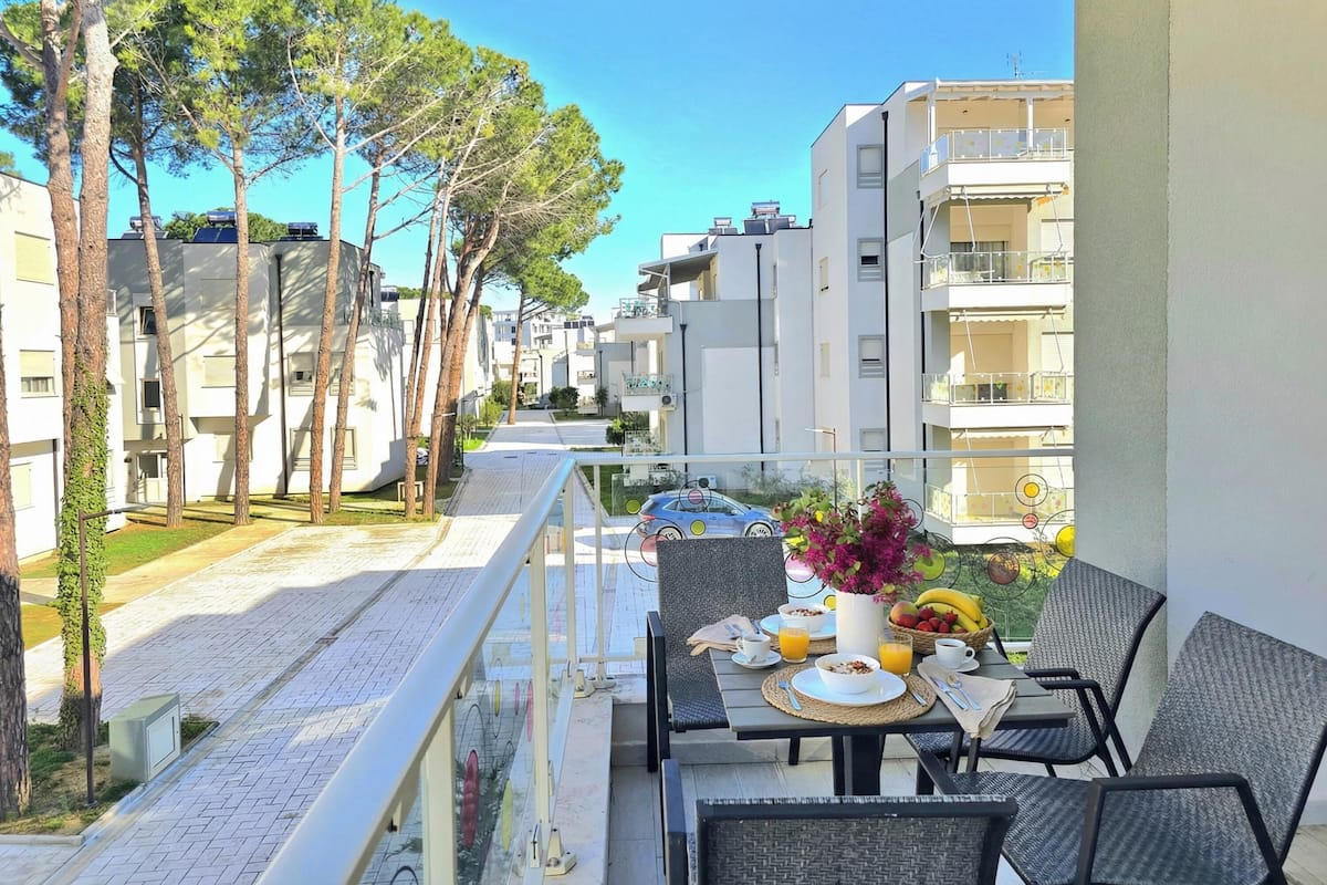 A balcony with outdoor seating, a table set with breakfast, and a view of a residential street with trees and buildings.