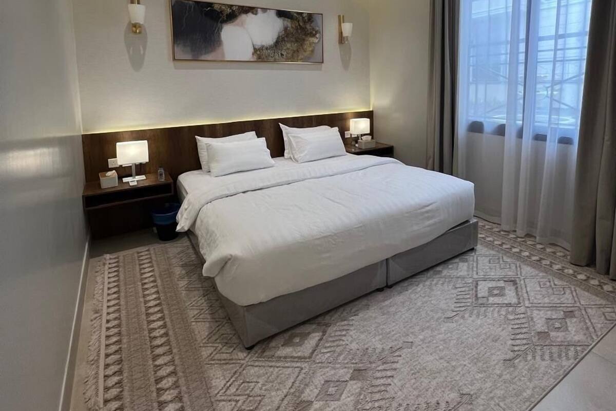 A modern hotel room with a large bed, a headboard with integrated lighting, a nightstand with a lamp, a wall-mounted artwork, and a view of the outdoors through sheer curtains.
