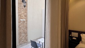 Une chambre d’hôtel comprenant un lit, une table de chevet, une lampe, une fenêtre avec des rideaux et une vue sur un espace extérieur aménagé avec une table et des chaises.