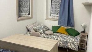 A small, neatly arranged room with a bed, a desk, and a chair. There are two windows with curtains, a lamp on a side table, and a small shelf. - Greta family house: Victory (Roma)