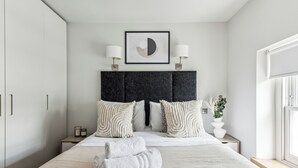 Un dormitorio moderno con una cama grande, un cabecero oscuro, dos lámparas de noche, una obra de arte enmarcada y una ventana con vistas.