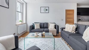 Una sala de estar moderna con un sofá gris, una mesa de centro de vidrio y una alfombra blanca. Se ve un ventanal con vistas a follaje verde.